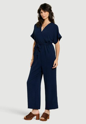 Vrouw draagt een marineblauwe wikkeljumpsuit met wijde pijpen en een tailleband, gecombineerd met bruine platformsandalen met hak.
