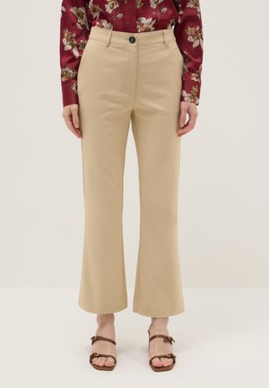 Frau trägt beigefarbene, hoch taillierte, leicht ausgestellt geschnittene Hose, kombiniert mit einer roten, langärmligen Bluse mit Blumenmuster und braunen Sandalen mit Absatz.