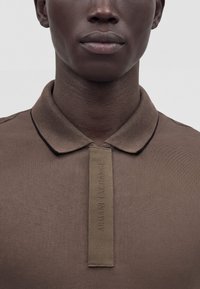 Κοντινό πλάνο ενός ατόμου που φορά ένα καφέ polo Armani Exchange με ταινία υφάσματος στη λωρίδα κουμπιών που δείχνει το όνομα της μάρκας.