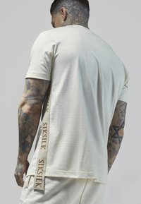 T-shirt a maniche corte di colore crema realizzata in un tessuto morbido, con una finitura testurizzata e un nastro con il logo laterale recante la scritta "SIKSIK" in tonalità tan.