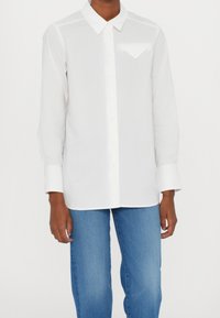 Weißes Langarmhemd mit Kragen, ausgestattet mit einer Brusttasche und Knopfleiste. Getragen mit blauen Jeans. Glatte Textiloberfläche.