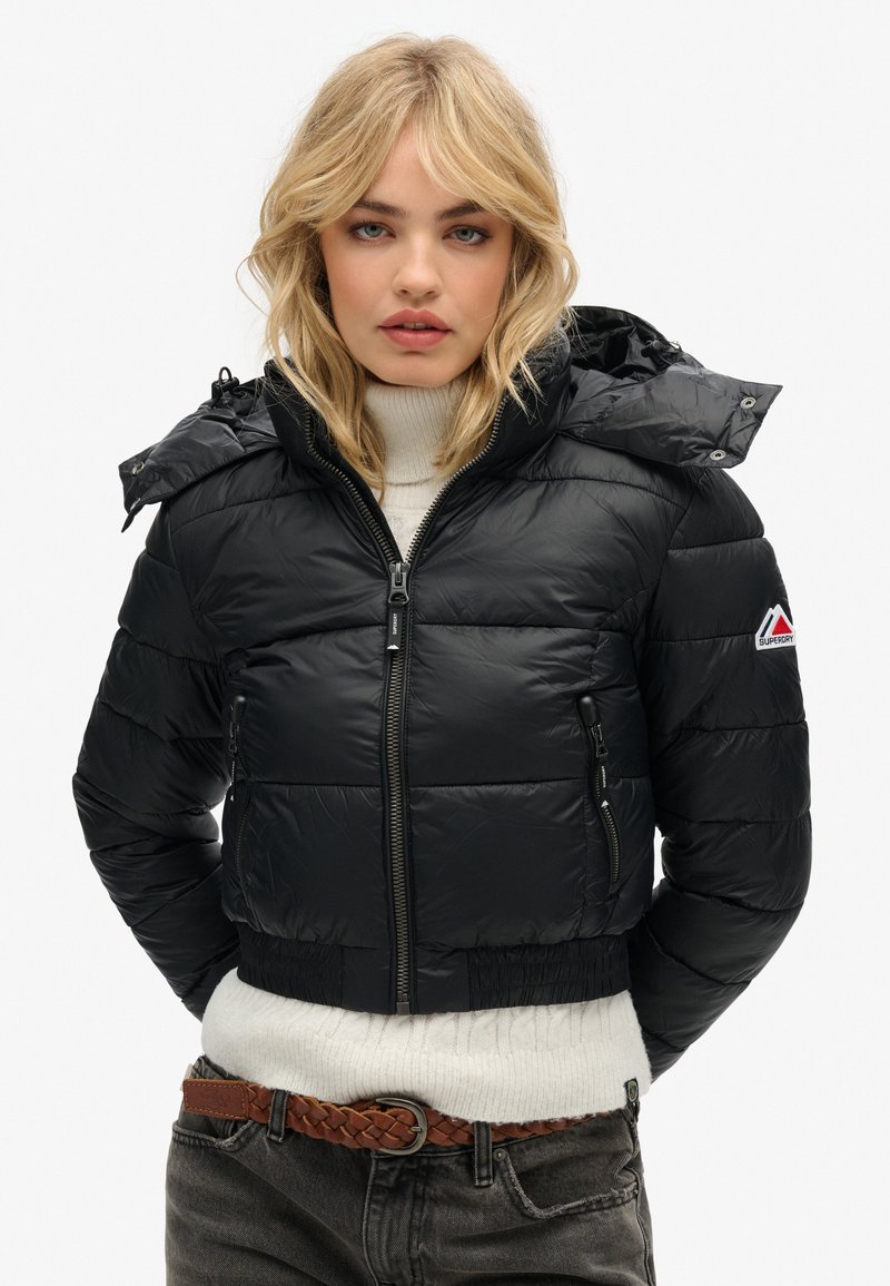 Giacca puffer nera corta con cappuccio; design trapuntato, chiusura frontale con zip, due tasche laterali e dettaglio del logo sul maniche. Indossata sopra un maglione crema.