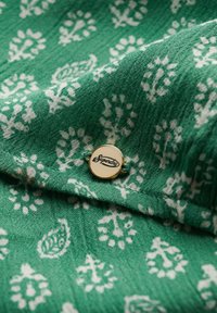 Tejido verde con un patrón floral blanco, que presenta un botón circular dorado con el texto "Superdry". Textura suave, detalle en capas.