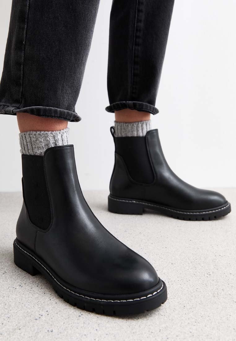 Bottes Chelsea en cuir noir avec des chaussettes en tricot gris et des pantalons noirs retroussés, posées sur un sol texturé gris clair.