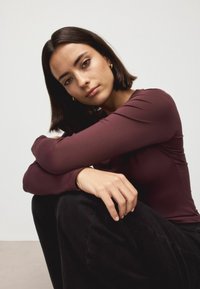 Burgunderfarbenes Langarmshirt mit glatter Textur, kombiniert mit schwarzen Hosen. Die Person hat schulterlanges dunkles Haar und sitzt mit auf den Knien ruhenden Armen.