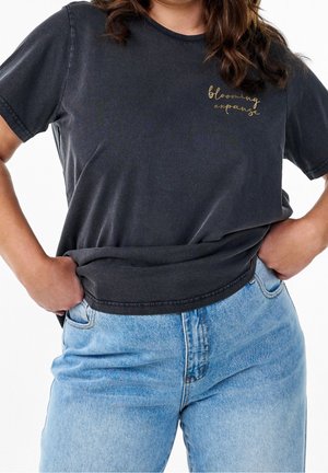 Kobieta w ciemnoszarej koszulce z napisem "blooming expanse" na piersi i jasnoniebieskich dżinsach, ręce wsunięte w przednie kieszenie.