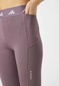 Violetie legingi ar platām elastīgām jostām, kas rotāti ar baltu logo. Iekļauj sānu kabatu un plakanas šuves komfortam.