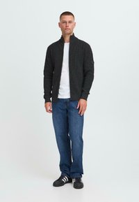 Uomo in piedi con una felpa zip-up nera, t-shirt bianca, jeans blu e scarpe da ginnastica nere con strisce bianche su sfondo chiaro uniforme.