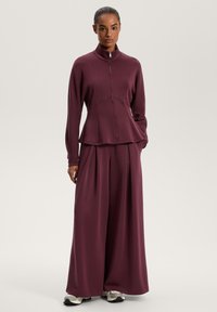Set sportiv bordo, format dintr-o jachetă cu fermoar și tiv peplum și pantaloni largi. Fabricat dintr-un material moale, cu cusături minime și un design sport.