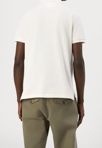 Homme portant un polo blanc uni à manches courtes et un pantalon vert olive, vu de dos sur un fond clair uni.