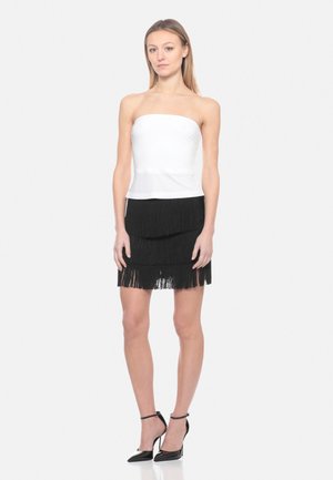 Donna che indossa un top bianco senza spalline, una minigonna nera a balze con frange e tacchi alti neri con cinturino alla caviglia, in piedi davanti a uno sfondo semplice.