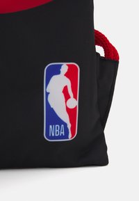 Geantă din țesătură neagră, cu un accent roșu și un logo rectangular NBA în albastru, roșu și alb, având silueta unui jucător de baschet.