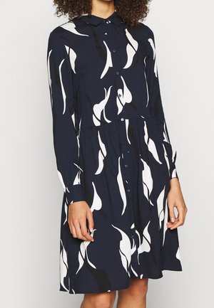 Femme portant une robe noire boutonnée avec des manches longues et des motifs abstraits de feuilles blanches.