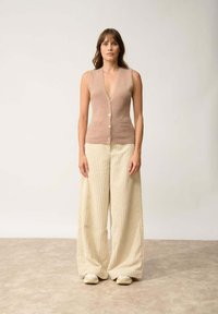 Beżowy wełniany bezrękawnik z głębokim dekoltem w kształcie V i guzikami z przodu, w połączeniu z kremowymi szerokimi spodniami z ryflowego materiału i jasnymi butami.