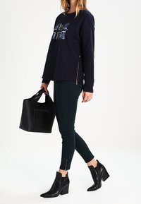 Mujer caminando, vistiendo una camiseta de manga larga azul marino con gráfico, pantalones oscuros ajustados con cremalleras en los tobillos, botas de tacón negras, llevando un bolso negro.