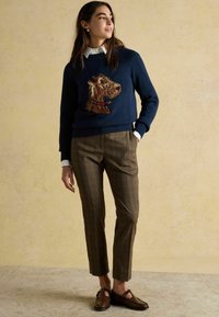 Tumes sinine sviiter, millel on koera motiiv ja ruuduline sall, on ühendatud pruuni ruudulise kolme-nelja pükstega. Pruun nahast loaferid täiustavad riietust.