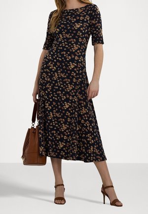 Femme portant une robe midi noire avec un petit motif floral marron, tenant un sac à main marron, assortie à des sandales marron à talons hauts.