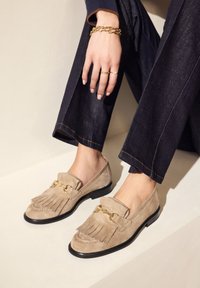 Beige suède loafers met leren franjes en gouden hardware, met een zwarte zool. Gepaard met donkere denim broek.