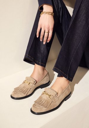 Beige suede loafers with leather fringe detail and gold hardware, featuring a black sole. Paired with dark denim pants.