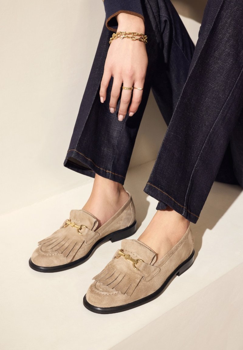 Beige suède loafers met leren franjes en gouden hardware, met een zwarte zool. Gepaard met donkere denim broek.