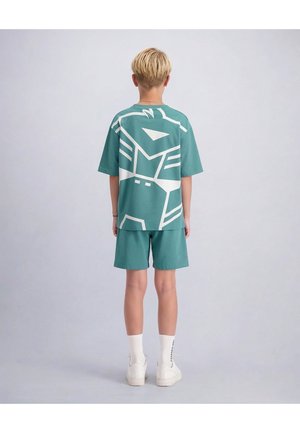Blond kind dat met zijn rug naar ons toe staat, draagt turquoise shorts en een oversized turquoise T-shirt met een groot wit abstract ontwerp op de rug.
