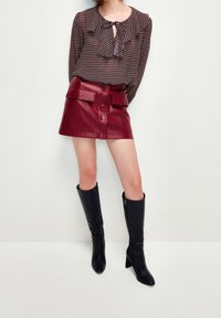Blusa marrón con estampado y cuello con volantes, combinada con una mini falda de piel sintética burdeos que tiene bolsillos y un cierre de botones en la parte delantera. Botas negras hasta la rodilla.