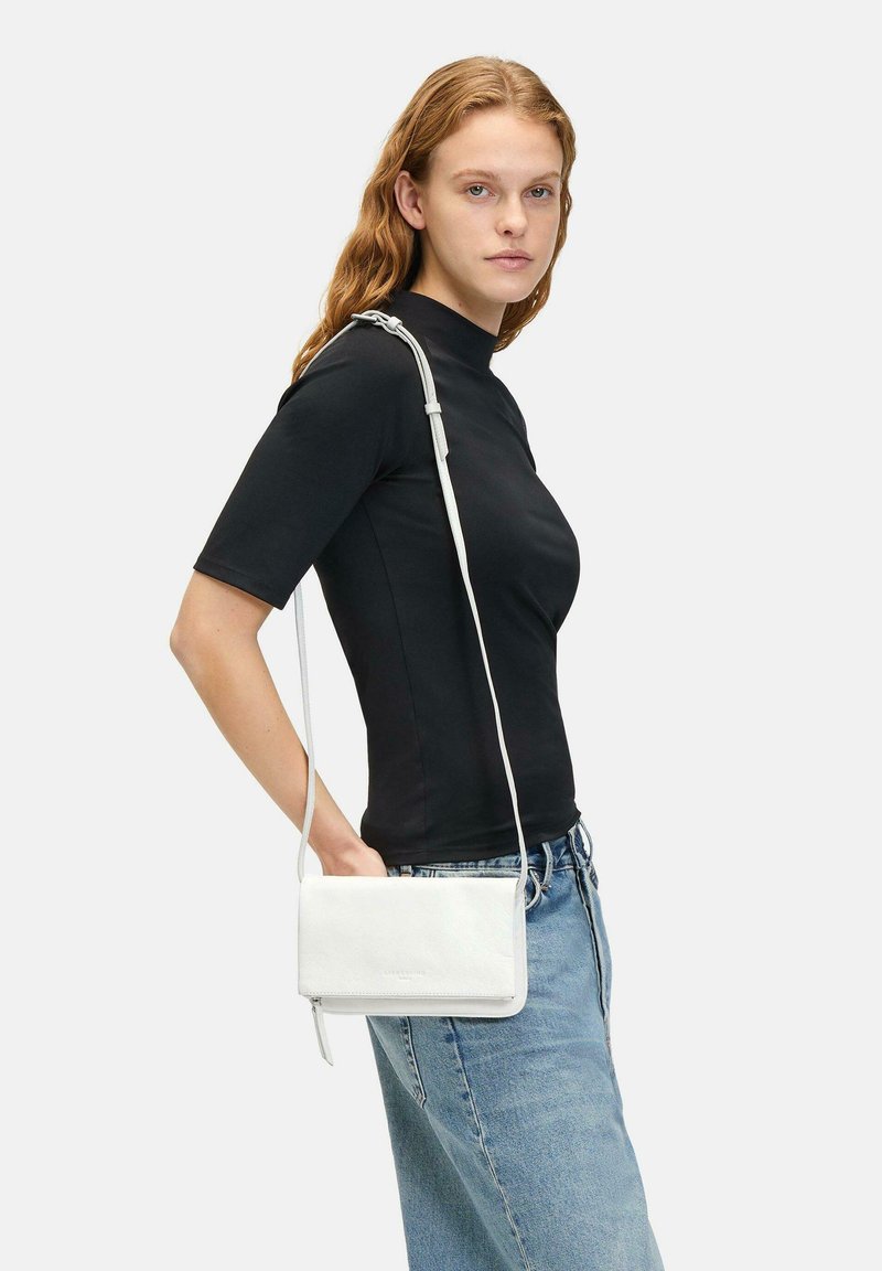 Biała skórzana torba na ramię z zamkiem błyskawicznym, z cienkim regulowanym paskiem, na tle modelki w czarnej bluzce i dżinsach.