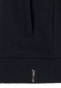 Tessuto blu navy scuro con una texture diagonale; presenta una tasca frontale e un'etichetta all'orlo. Design classico, adatto per un abbigliamento casual.