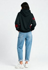 Sudadera negra con capucha y patrones de corazones rojos en las mangas, combinada con unos jeans azul claro con puños y zapatillas blancas. Textura de tela suave.