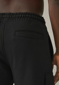 Los pantalones de chándal negros cuentan con una cinturilla elástica texturizada, un bolsillo lateral y una superficie de tela suave. El diseño es sencillo y funcional.