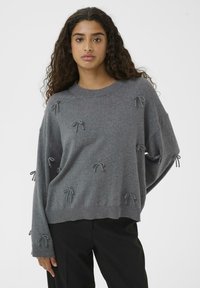 Gray knit sweater featuring loose-fitting design, round neckline, and textured accents with decorative bows. Paired with black pants.