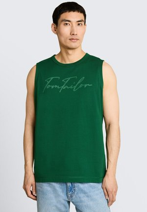 Man draagt een mouwloos donkergroen shirt met het "Tom Tailor" logo, gecombineerd met lichtblauwe spijkerbroek, staand tegen een effen grijze achtergrond.