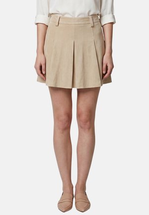 Femme portant une mini-jupe plissée beige, une chemise blanche à manches longues avec manches retroussées et des ballerines nude à bout pointu, debout avec les mains le long du corps.