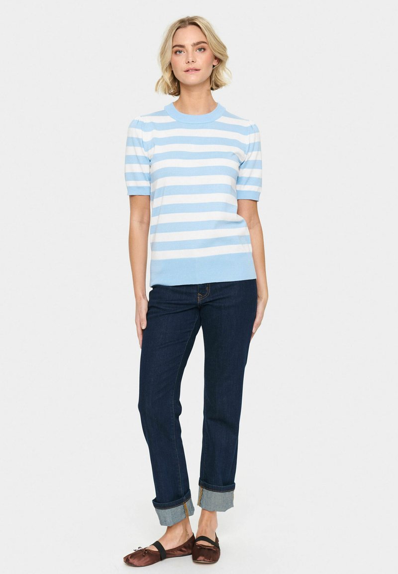 Jasnoniebieski i biały sweter dzianinowy w paski z krótkimi bufiastymi rękawami, w połączeniu z ciemnoniebieskimi dżinsami podwiniętymi na mankietach. Brązowe płaskie buty.