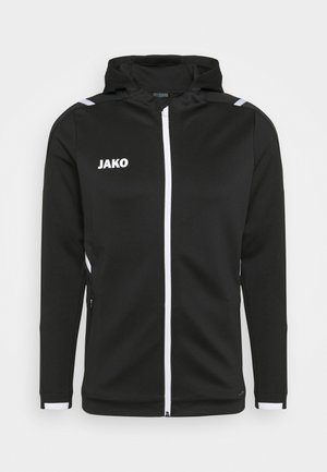 Veste noire à capuche en tissu texturé, avec une fermeture éclair blanche et des accents, portant le logo "JAKO" sur la poitrine gauche et des poches latérales.