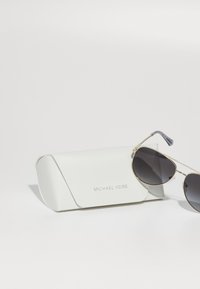 Guld aviator-solbriller med mørke linser ved siden af en hvid tekstureret etui, der har det præget "MICHAEL KORS" logo på forsiden.