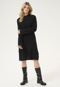 Abito nero a maglia con maniche lunghe, texture a coste e spacchi laterali. Abbinato a stivali neri lucidi fino al ginocchio con dettagli in fibbia.