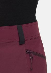 Bordeauxkleurige broek met een gladde textuur, voorzien van een zwarte tab aan de tailleband en een ritszakdetail aan de zijkant.