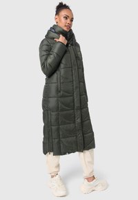 Langdonkergroene pufferjas met een hoge kraag, rits- en drukknopensluitingen, zijzakken en een quiltpatroon. Beigekleurige broek en schoenen maken de look compleet.