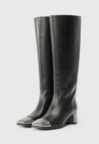 Mustad nahast põlvekotid, mille esiosal on ümarad hõbedased nupud ja plokkninad, seisab sirgelt ühetoonilise heleda taustal.