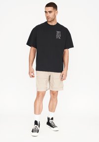 Camiseta negra de gran tamaño con un estampado gráfico en el lado izquierdo del pecho, combinada con pantalones cortos de cargo beige y zapatillas altas negras. Calcetines blancos visibles.