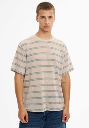 Joven con cabello corto que lleva una camisa beige de manga corta con rayas horizontales azul claro y vaqueros azules, de pie frente a un fondo blanco.