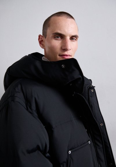 Junger Mann mit short Haar, der eine schwarze Daunenjacke trägt und selbstbewusst in die Kamera schaut, vor einem schlichten grauen Hintergrund.