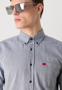 Cămașă gri cu nasturi, cu o textură specială, având un buzunar pe piept și un mic logo roșu, însoțită de ochelari de soare rectangulari și transparenți.