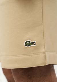 Μπεζ σορτς με μικρό πράσινο κεντημένο λογότυπο κροκόδειλου κοντά στο τελείωμα, φορεμένα από άτομο με σκουρόχρωμο δέρμα.