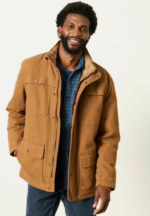 Mann lächelt, trägt eine braune Jacke mit Schafsfellkragen über einem blau karierten Hemd und dunklen Jeans, steht vor einem schlichten Hintergrund.