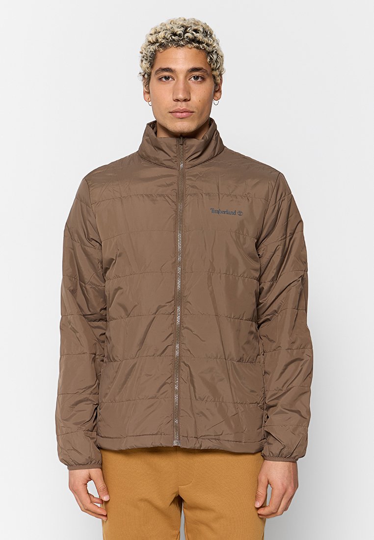 Giacca imbottita marrone con colletto alto e chiusura a zip. Presenta un logo sottile e cuciture orizzontali. Tessuto leggero e testurizzato.
