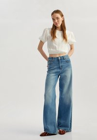 Hvid cropped bluse med puffærmer, parret med lys blå brede jeans. Brune sko har guldnitter som accenter. Neutral baggrund.