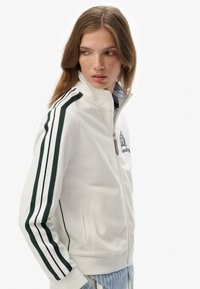 Hvid atletisk jakke med høj krave, grønne sideløsninger, broderet logo og lynlåsefront. Har sidelommer og en blød tekstur.