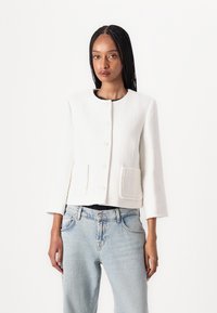 Weißer strukturierter Blazer mit sechs Knöpfen, runder Halslinie und zwei vorderen Taschen. Getragen über hellblauen Jeans.